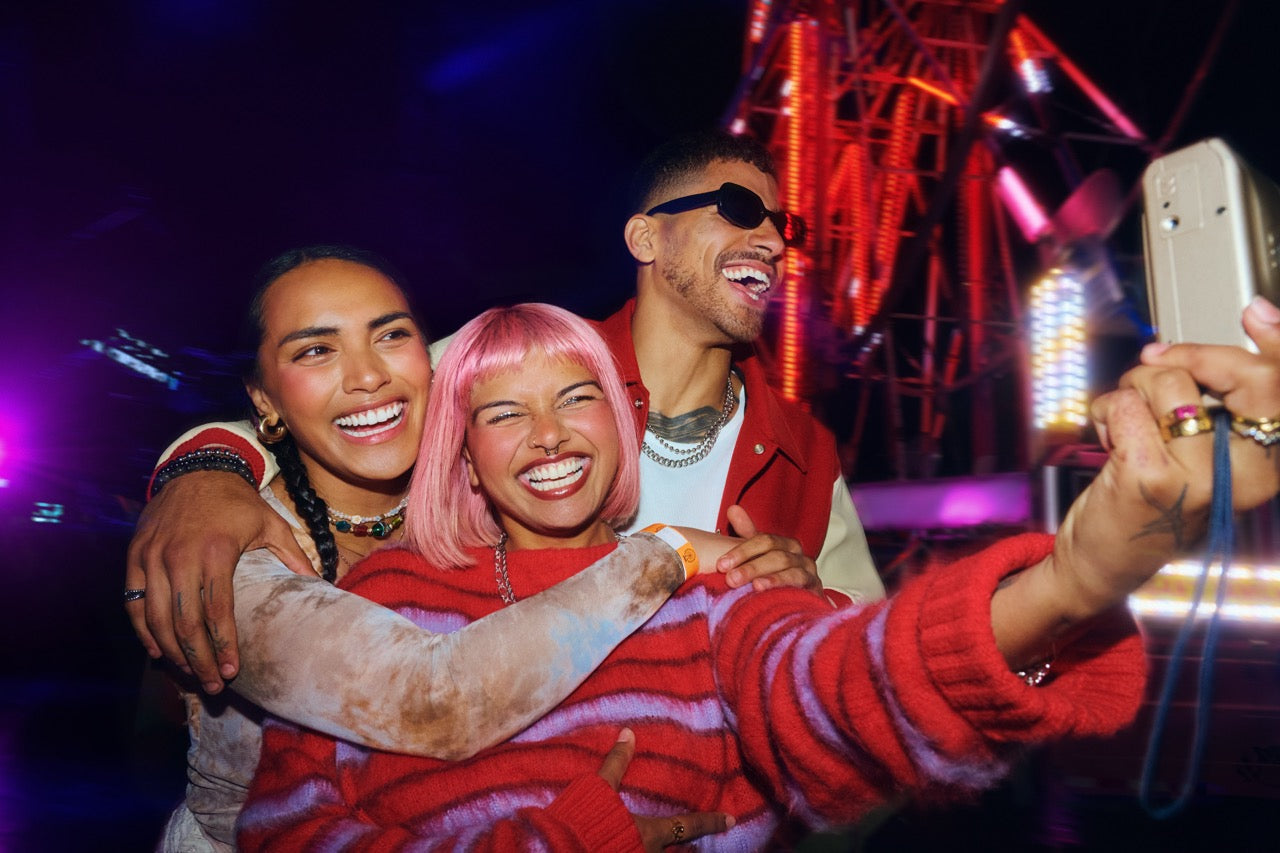 Three friends laughing and taking a selfie at a vibrant nighttime event with colorful lights